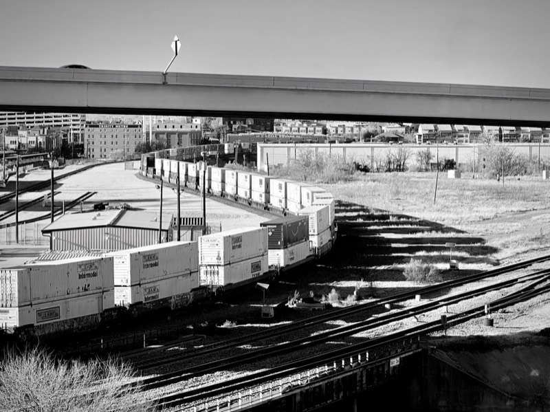 A freight train and stacked containers in a black and white rail yard photograph.